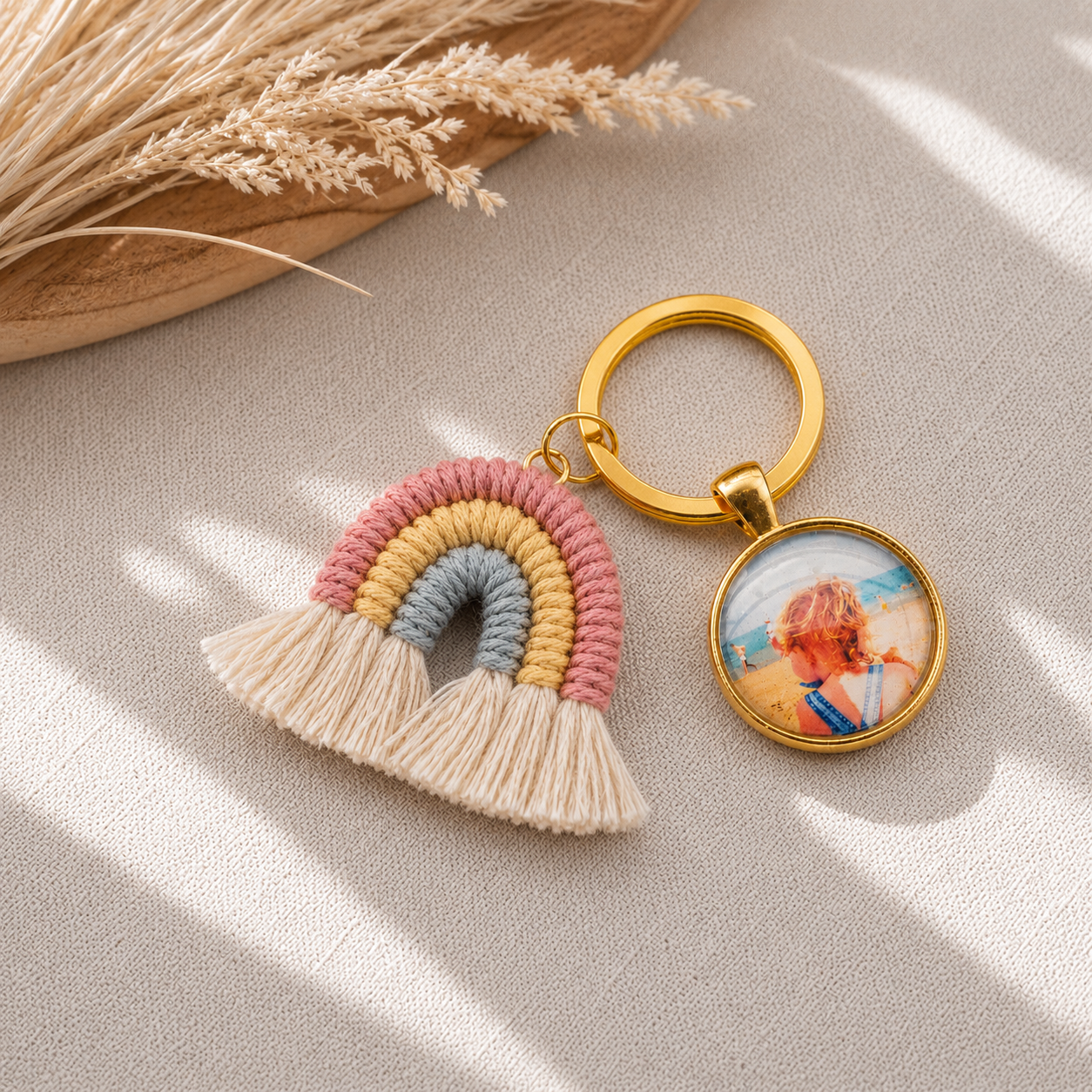 Personalisierter Makramee-Schlüsselanhänger mit Regenbogen-Anhänger in Rosa, Beige und Hellblau sowie rundem Fotoanhänger in Goldoptik auf hellem Stoff im Boho-Stil.