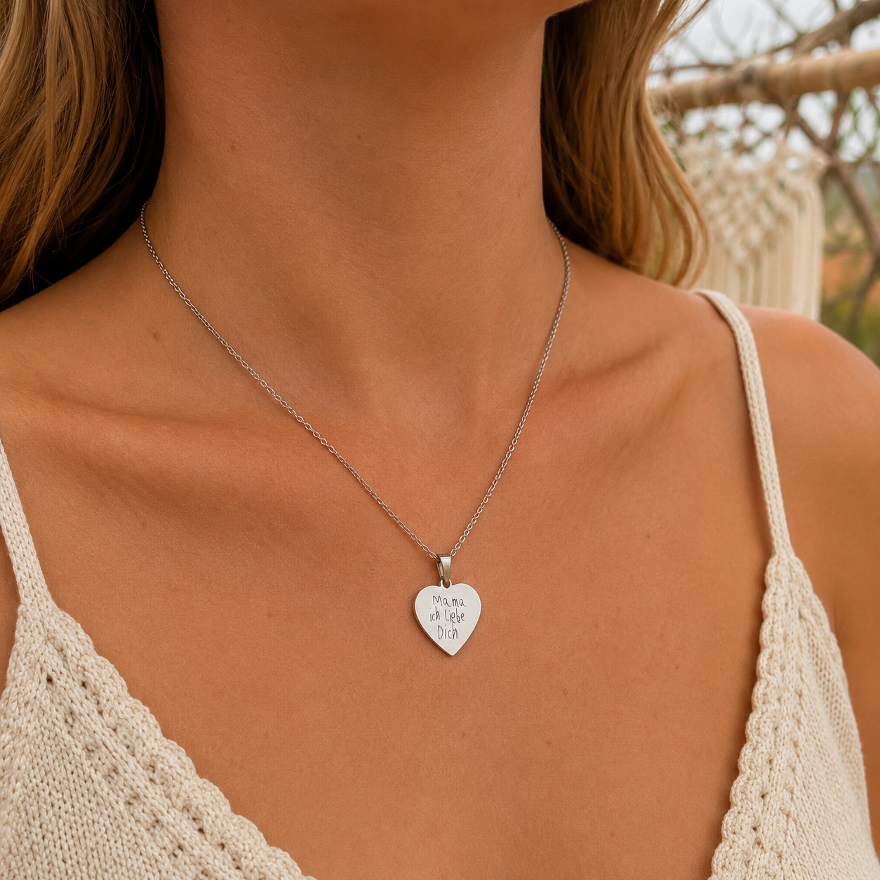 Nahaufnahme einer Frau mit einer silberfarbenen Halskette und herzförmigem Anhänger, graviert mit der originalen Kinderhandschrift „Mama ich liebe Dich“, vor einem warmen Boho-Hintergrund.