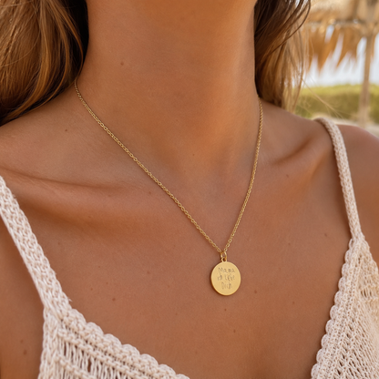 Frau trägt eine goldfarbene Halskette mit rundem Gravuranhänger und originaler Kinderhandschrift „Mama ich liebe Dich“ vor einem sommerlichen Boho-Hintergrund.