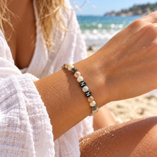 Personalisiertes Natursteinarmband mit cremefarbenen, beigen, grauen und braunen Perlen sowie zwei schwarzen Acrylperlen mit weißen Wunschbuchstaben. Das Armband wird am Handgelenk einer Frau vor sommerlichem Strandhintergrund getragen und ist in verschiedenen Farbvarianten und Wunschgrößen erhältlich.