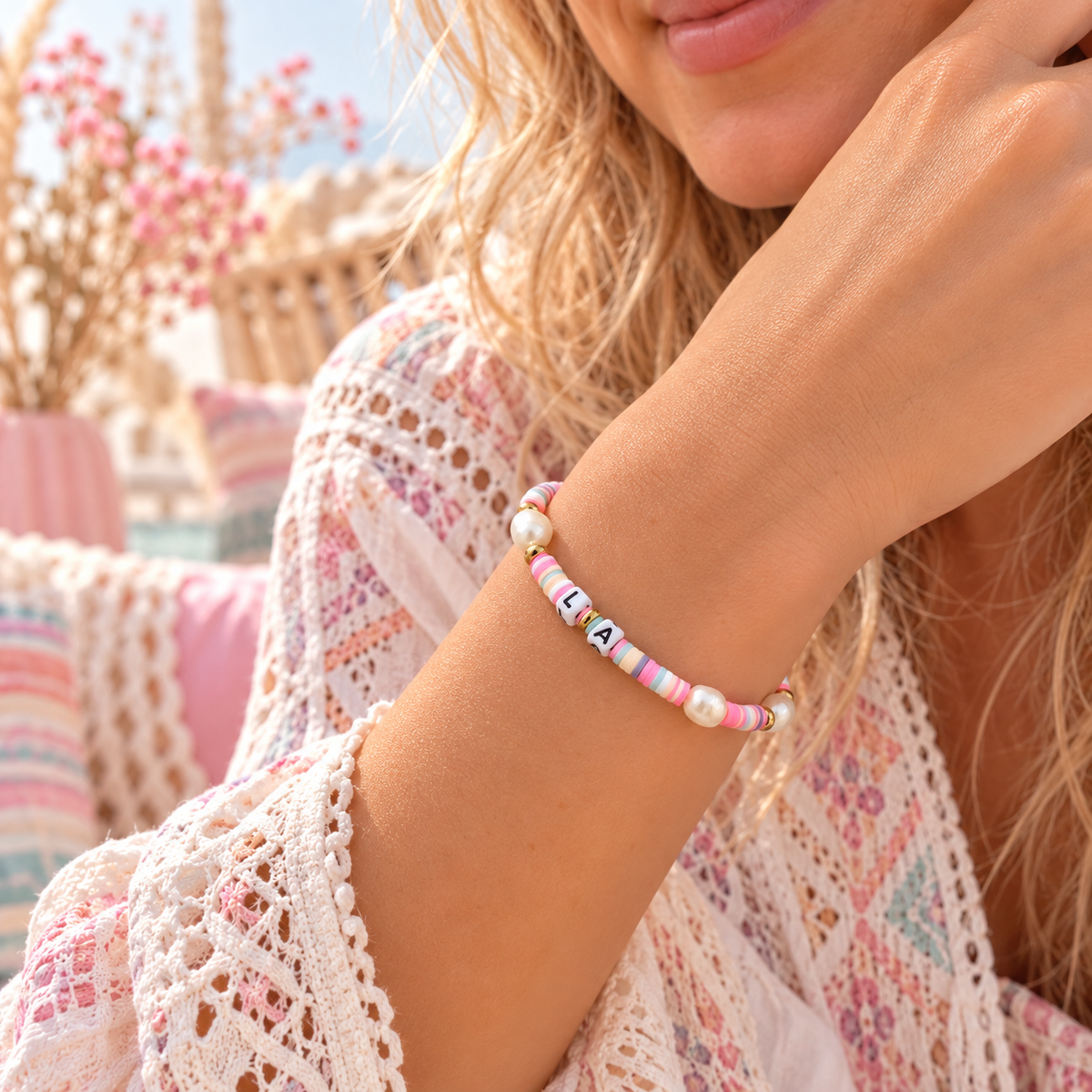 Nahaufnahme eines zarten, personalisierbaren Damenarmbands am Handgelenk einer Frau. Das Armband besteht aus bunten Polymerperlen in Pastelltönen, echten Süßwasserperlen, goldfarbenen Edelstahlperlen und Buchstabenperlen. Romantischer Boho-Look mit sanftem, pastellfarbenem Hintergrund.
