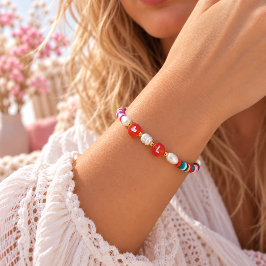 Farbenfrohes, personalisiertes Damenarmband mit bunten Polymerperlen, echten Süßwasserperlen, goldfarbenen Edelstahlperlen, roter Herzperle und Buchstabenperle mit L. Getragen am Handgelenk einer Frau vor einem zarten, romantischen Boho-Hintergrund.